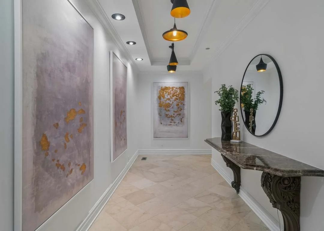 A hallway featuring three pink and gold abstract paintings on the left wall, a round mirror on the right wall, a marble console with potted plants, a decorative sculpture, and three black and gold pendant lights hanging from the ceiling.