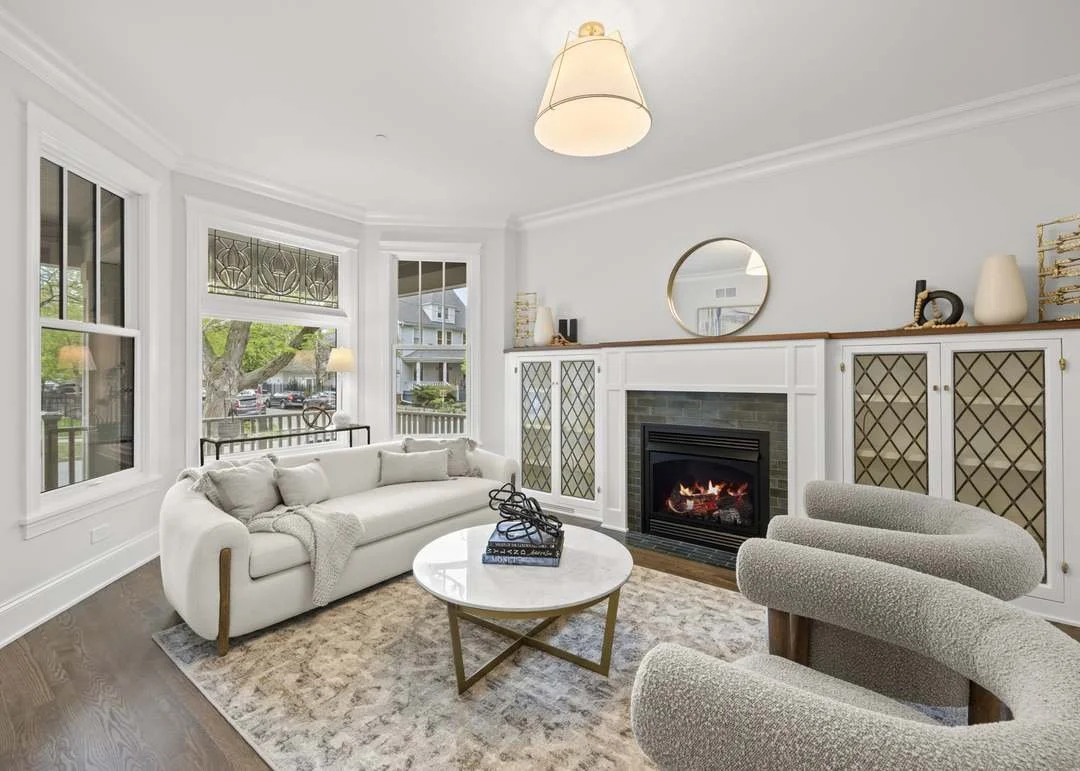 Living room with a white sofa, round coffee table, fireplace, and upholstered armchair, with large windows showing trees and houses outside.