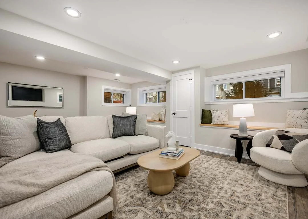 A modern living room with a white sofa, dark and light throw pillows, a beige armchair, a wooden coffee table, a patterned area rug, multiple windows, and a white door.