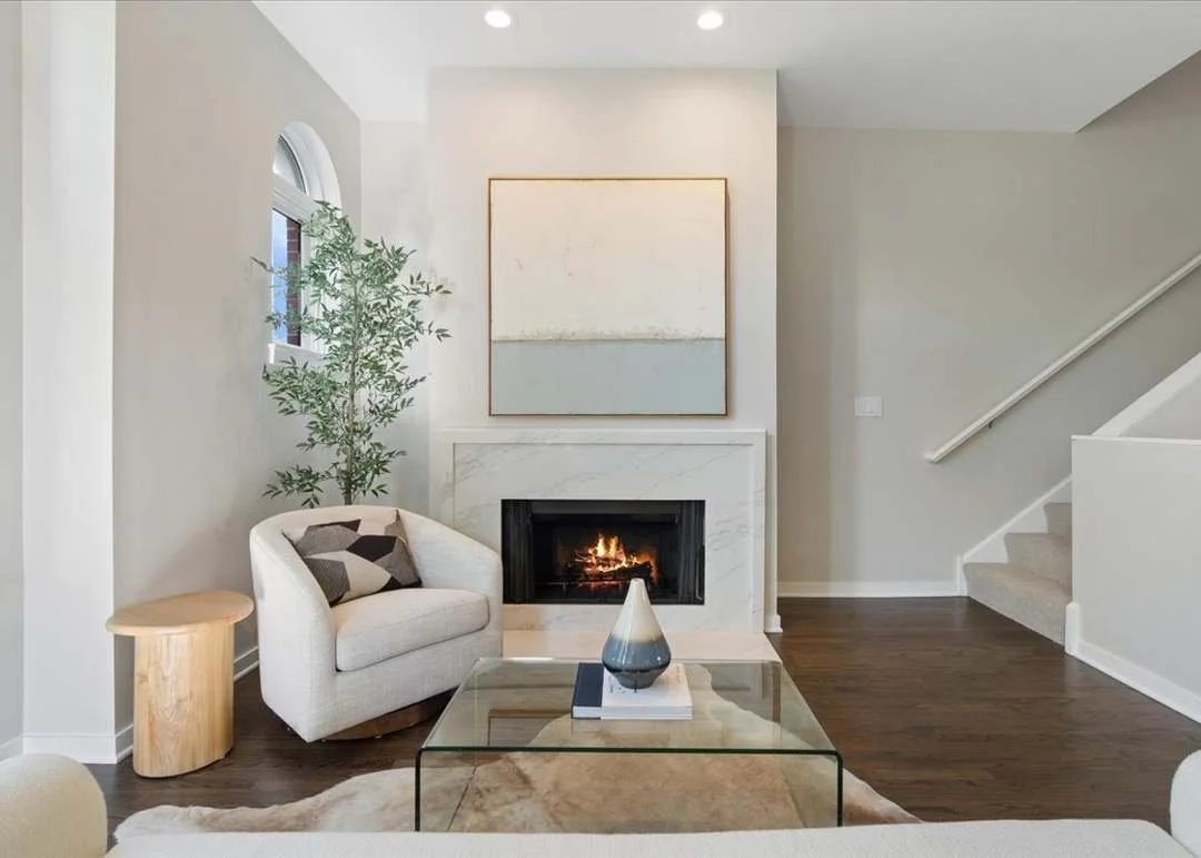 Living room with a fireplace, a white armchair with a gray pillow, a glass coffee table with a decorative vase, a small wooden side table, a tall green plant, a large abstract painting above the fireplace, and hardwood floors.