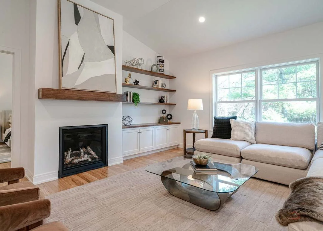 Living room with a beige sectional sofa, glass coffee table, wooden shelves with decorative items, tall window with trees outside, white walls, hardwood floors, and a fireplace.