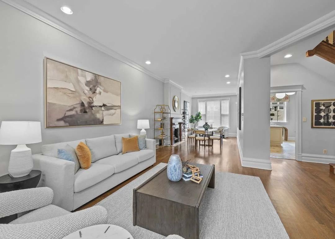 A spacious living room with a white sofa, colorful pillows, a coffee table, and a large abstract painting on the wall. There is a dining area with a round table and chairs in the background, along with windows bringing in natural light.