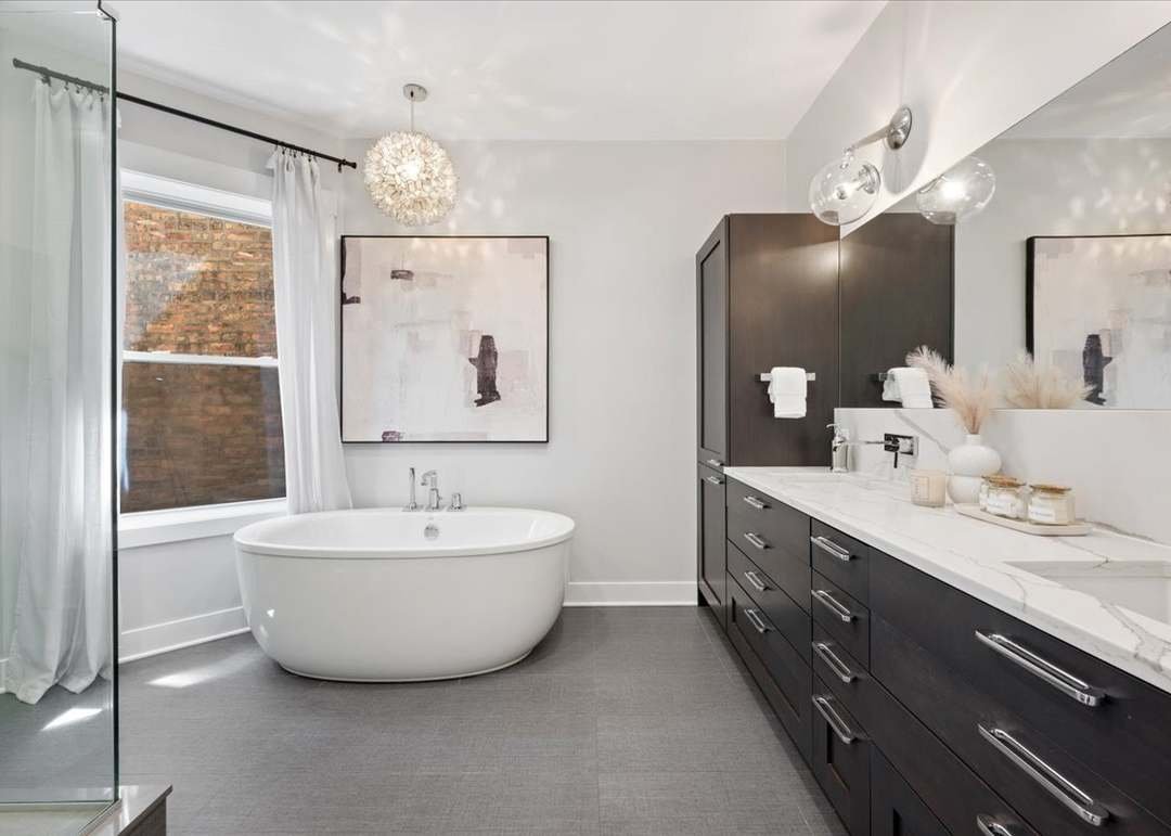 Modern bathroom with a white freestanding bathtub, large window with white curtains, dark wood cabinetry with a marble countertop, and abstract artwork on the wall.