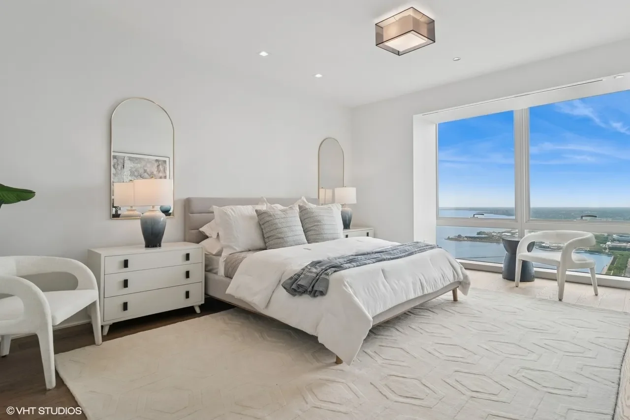 A modern bedroom with white walls, a large bed with white bedding and gray pillows, a white rug, two nightstands with lamps, a wooden chair, and a large window showing a cityscape and water view.