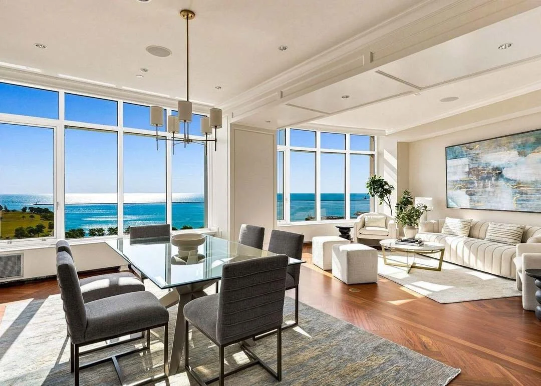 Living and dining room with large windows overlooking the ocean, modern furniture, a glass dining table with six chairs, and a white sofa with artwork on the wall.