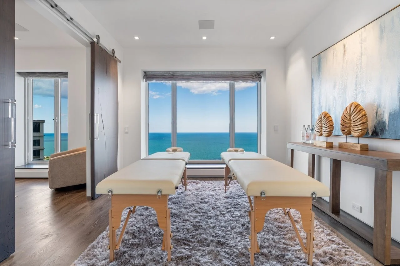 A massage room with two massage tables facing a large window showing an ocean view, a wooden sideboard with decorative leaves and bottles, and a shaggy rug on wooden floors.