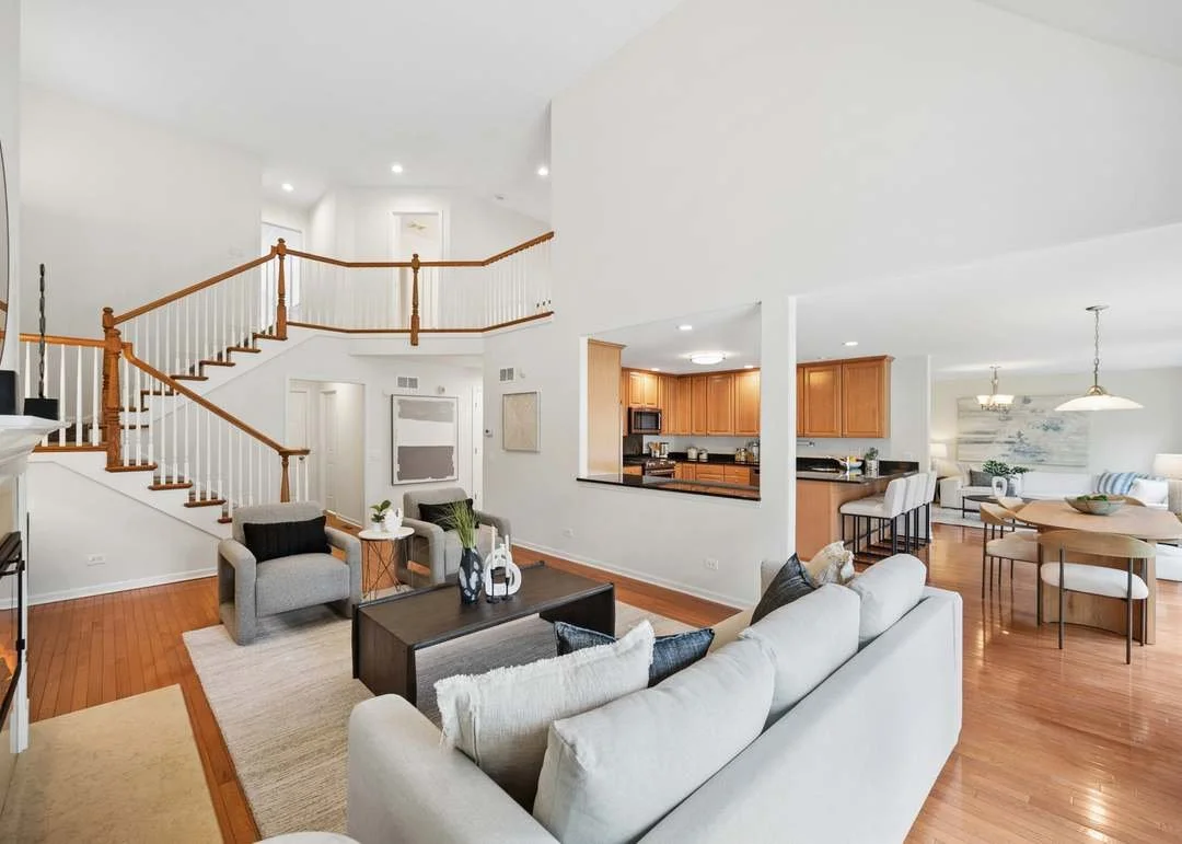 Open-concept living room with a white sofa, armchairs, a coffee table, and a staircase with wooden handrails leading to the second floor. Adjacent to the living area is a kitchen with wooden cabinets and an adjacent dining area with a round table and chairs.