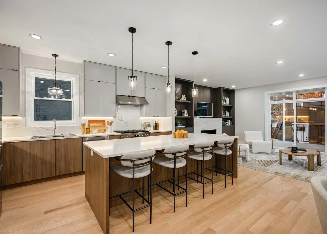 Open-concept kitchen and living room with modern design, wooden flooring, white and gray cabinetry, kitchen island with bar stools, and a sitting area with armchairs and a TV