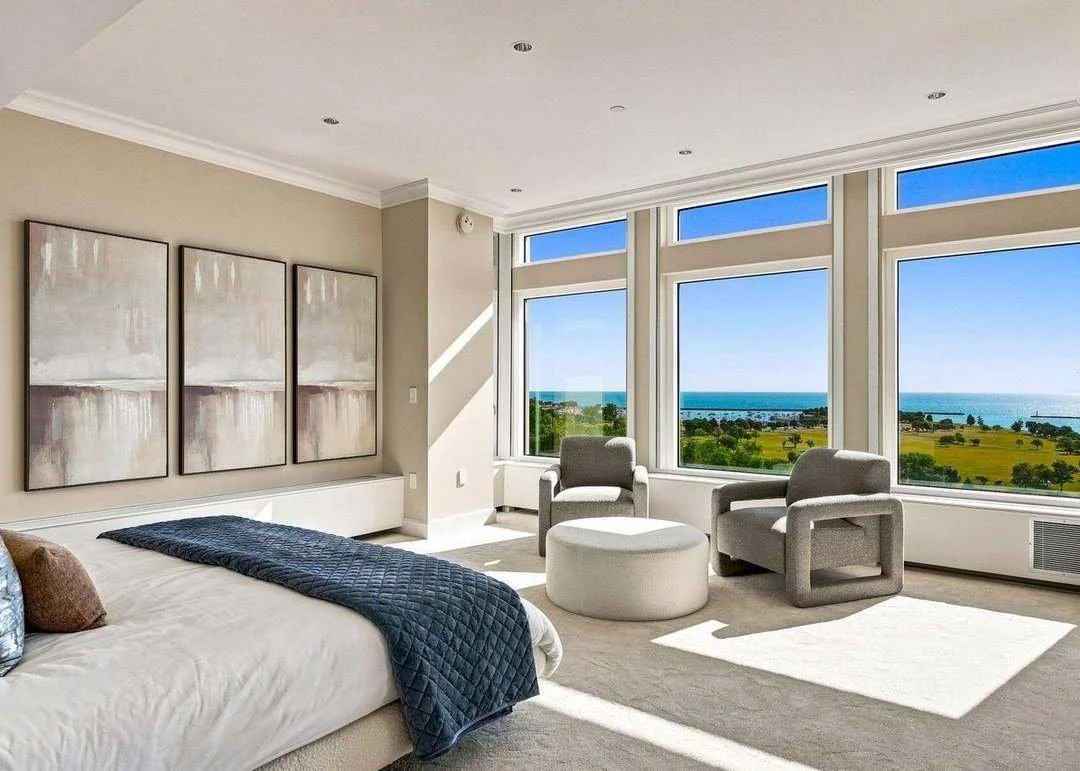A bedroom with large windows overlooking a green landscape and ocean, with two gray armchairs, a white ottoman, and abstract wall art.