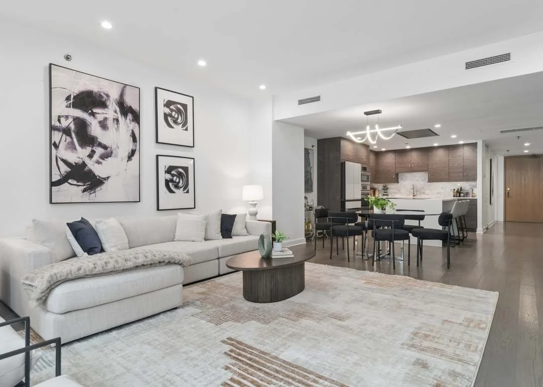 Modern living room and kitchen with white walls, dark wood flooring, and contemporary furniture. The living room has a white couch with black and white pillows, abstract art on the wall, and a round coffee table. The kitchen features a dining table, black chairs, and a white island with bar stools.