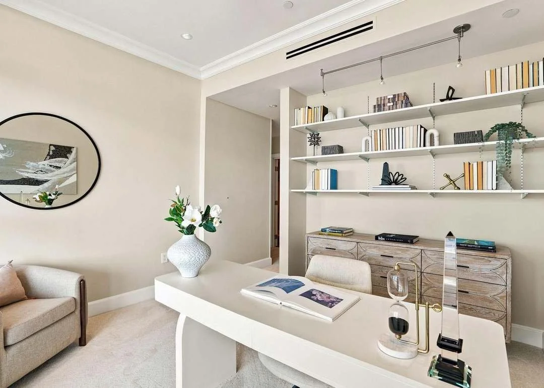 A modern office space with a white desk, a beige sofa, and a wall-mounted bookshelf filled with books and decorative items. There is a large mirror on the wall and a white vase with flowers on the desk.