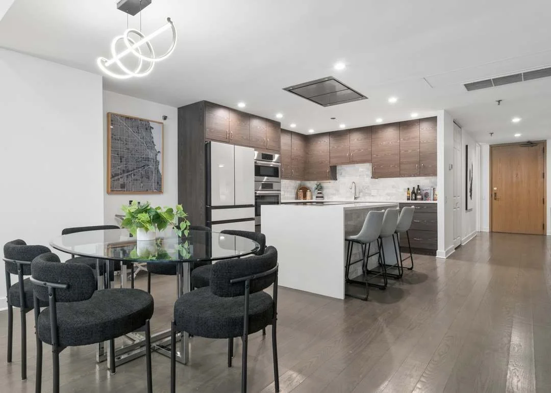 Modern open-concept kitchen and dining area with a round black table, six dark gray chairs, a white kitchen island with barstools, dark wood cabinets, stainless steel appliances, and a decorative light fixture.