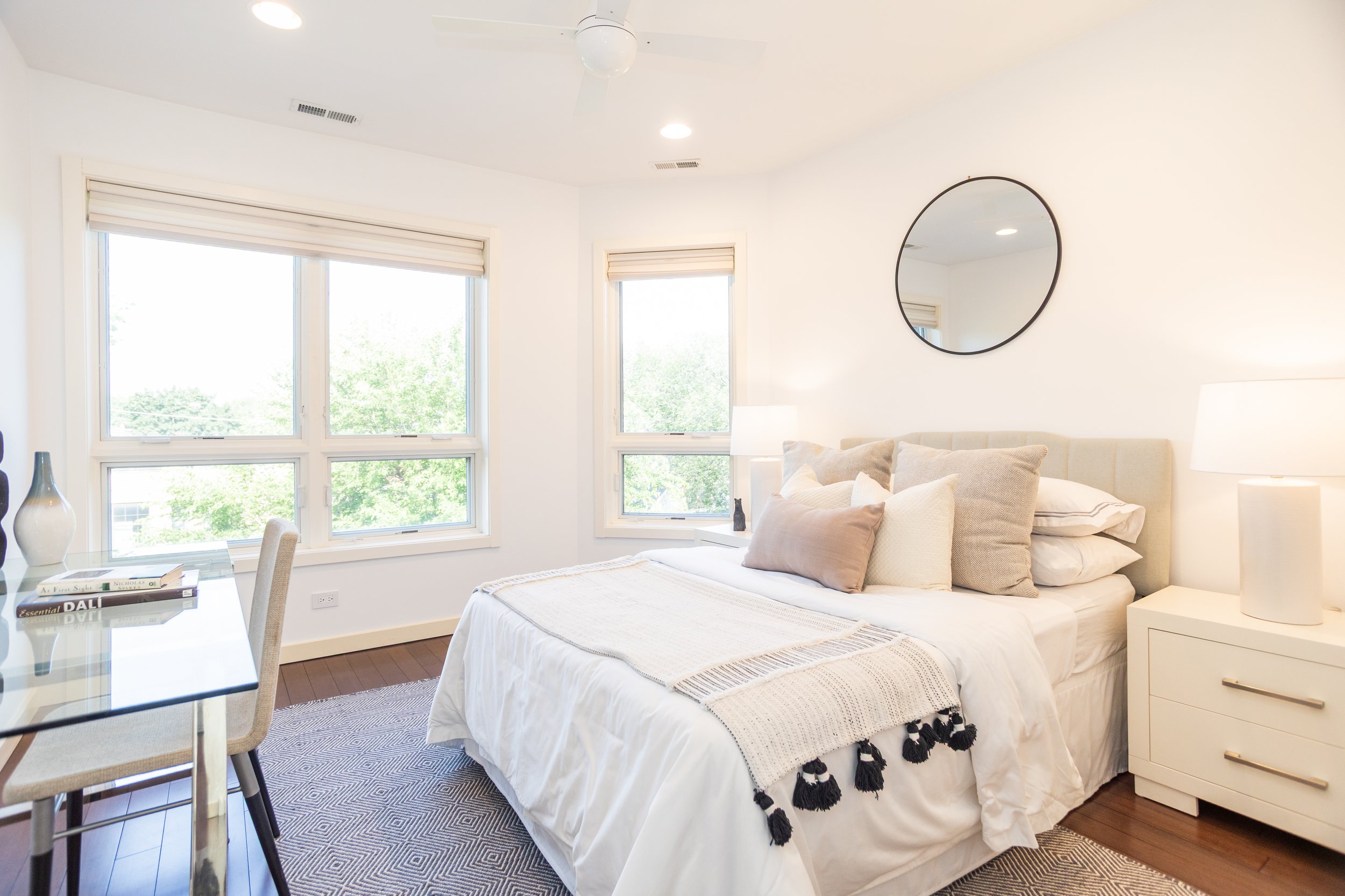 Bright bedroom with a large bed, multiple pillows, white bedside tables with lamps, three windows with blinds showing greenery outside, a mirror on the wall, and a glass table with books and vases.