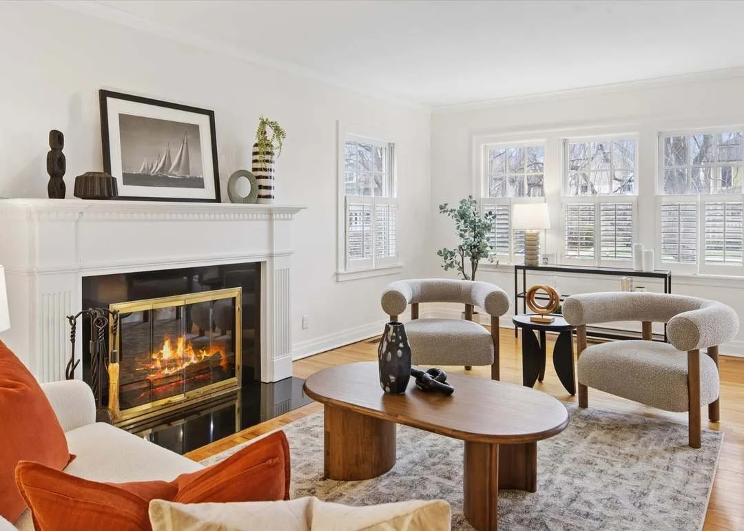 Living room with a fireplace, modern art and decorative vases on mantel, beige armchairs, a coffee table with vases and shoes, large windows with shutters, and a potted plant.