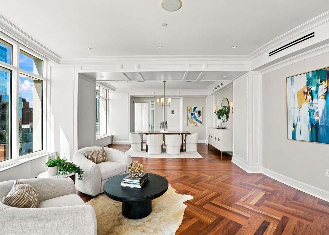 Modern living room with white walls, wooden floors, large windows, and minimal decor. Contains beige sofas, a black coffee table, a white sideboard, and colorful abstract artwork.