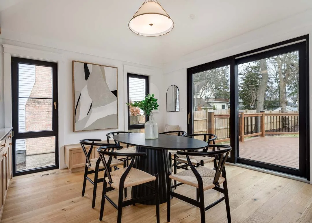 Dining room with a black oval table, six chairs with woven seats, a vase with green foliage, wall art, a mirror, large sliding glass door leading to a deck, and two windows showing trees and houses outside.