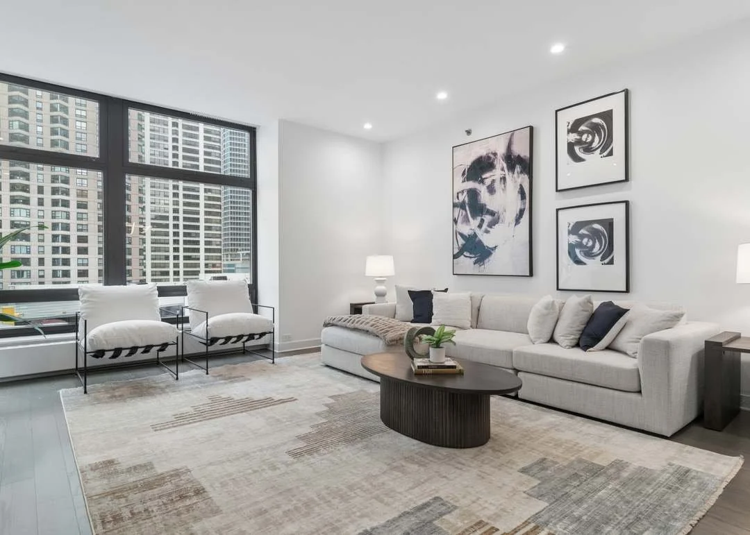Modern living room with large window showing city buildings, white sofa with pillows, two white armchairs, abstract artwork on the wall, coffee table with decor, and lamps.