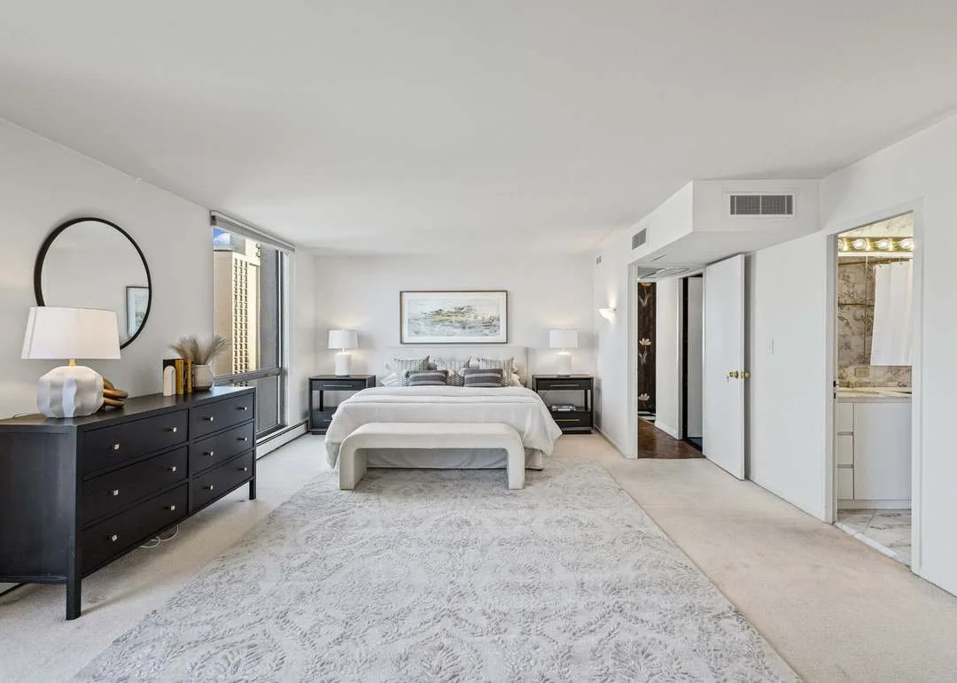 Modern bedroom with white walls and bedding, black nightstands, a black dresser with a round mirror, and a large area rug. There is a window with light gray curtains and a framed picture above the bed. Two lamps are on the nightstands.
