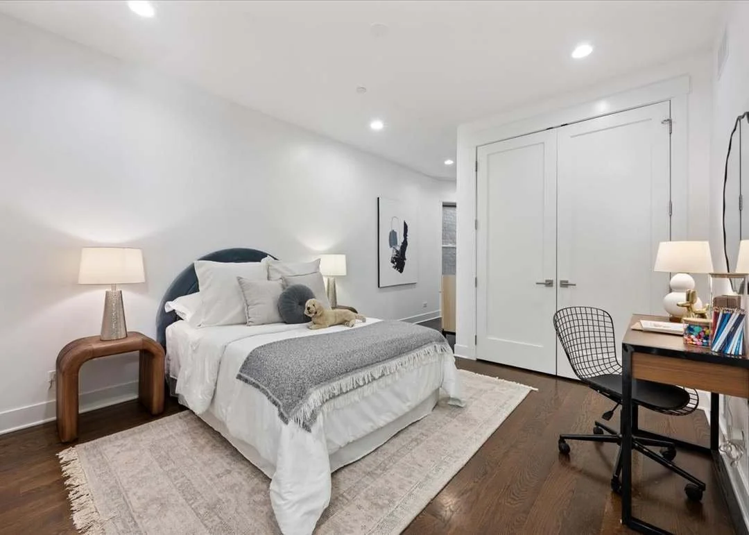 A modern bedroom with white walls, hardwood flooring, and a bed with white and gray bedding. There are two bedside lamps, a small dog lying on the bed, and a framed art piece on the wall. A wooden nightstand, a black wire chair, and a desk with stationery are also visible.
