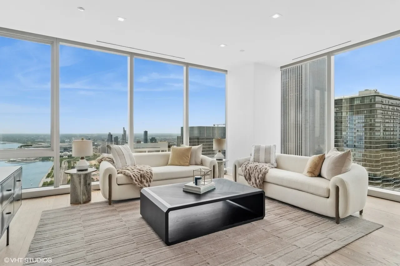 A modern living room with two white sofas, a black coffee table, and large floor-to-ceiling windows showing a city skyline and river view.