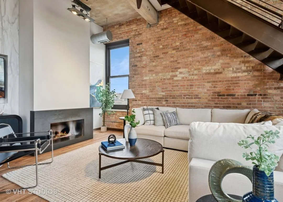 Modern living room with exposed brick wall, a white sectional sofa, a black round coffee table, a beige area rug, and a fireplace. There is a window with a view of the sky, a floor lamp next to the sofa, indoor plants, and a staircase on the right.