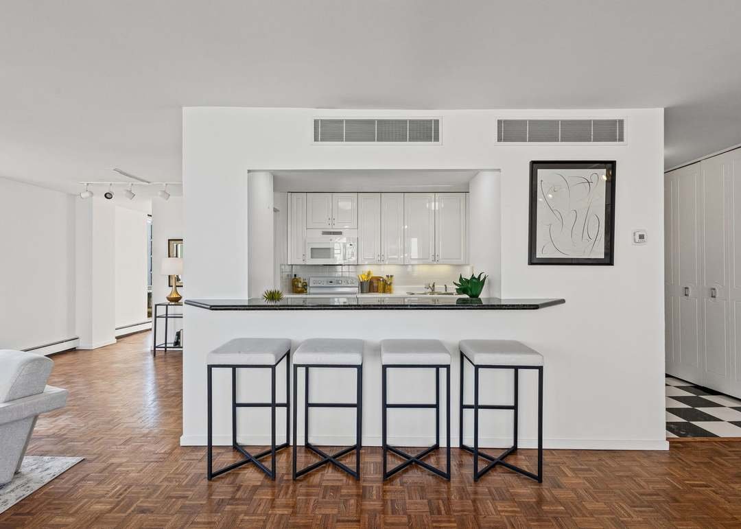 Kitchen with white cabinets, microwave, and countertop with bar stools in front, hardwood floor, living room on the left, and hallway on the right, minimalistic decor with framed artwork on the wall.
