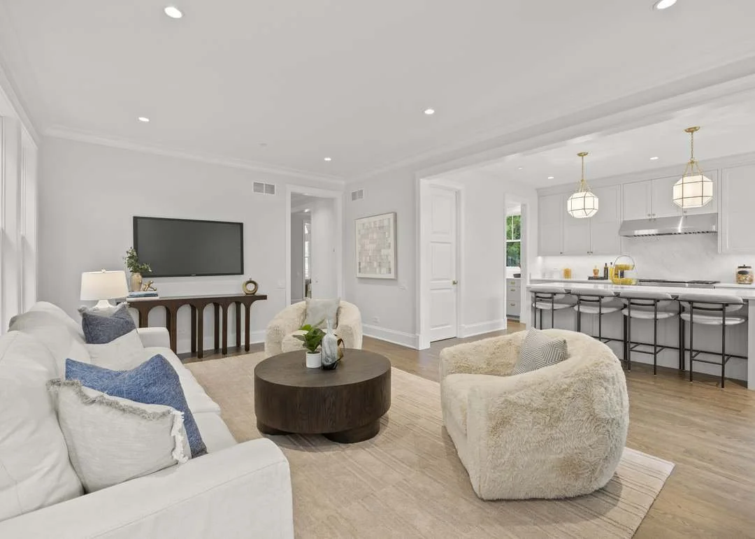 Living room with white sofa and plush armchairs, coffee table, wall-mounted TV, and view of modern kitchen with bar stools and pendant lights.