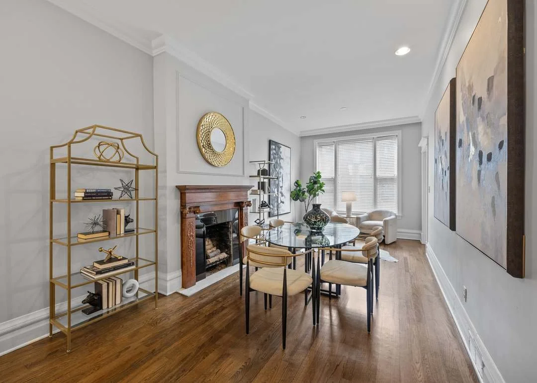 Living room with wooden floor, white walls, a fireplace with a wooden mantel, modern artwork, a round glass dining table with six chairs, gold shelving with decorative items, large windows with blinds, and a white armchair near the window.