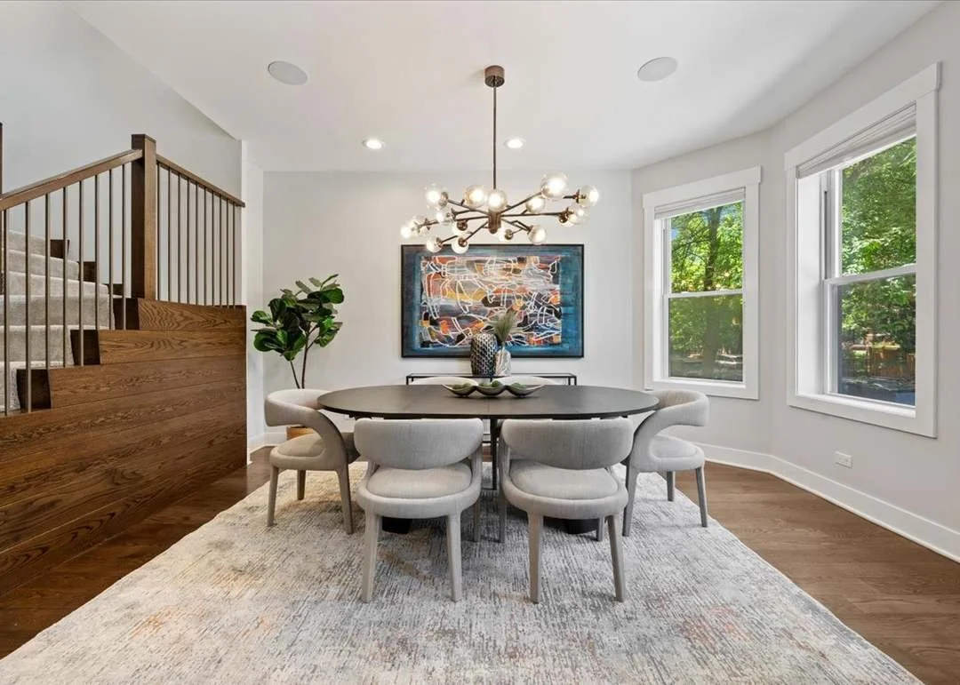 Modern dining room with a round black table, beige upholstered chairs, a chandelier, a framed abstract artwork on the wall, a potted plant, large windows showing green trees, and hardwood floors with a light-colored rug.