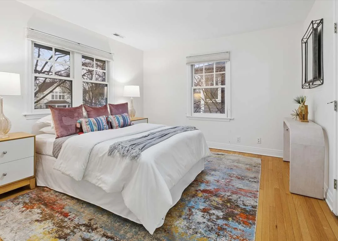 A bedroom with white walls, a wood floor, and three windows. The bed has white bedding, pink pillows, and a colorful bolster pillow. There are two white nightstands with lamps, a rug with multicolored abstract patterns, and a small white dresser with a plant and a decorative piece. A black mirror hangs on the wall.