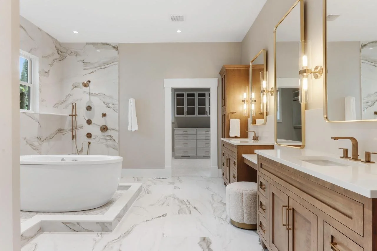 A modern bathroom with a white marble floor and backsplash, a freestanding bathtub, a walk-in shower with a window, wooden vanity with dual sinks, large mirrors with gold frames, and a small round upholstered stool.