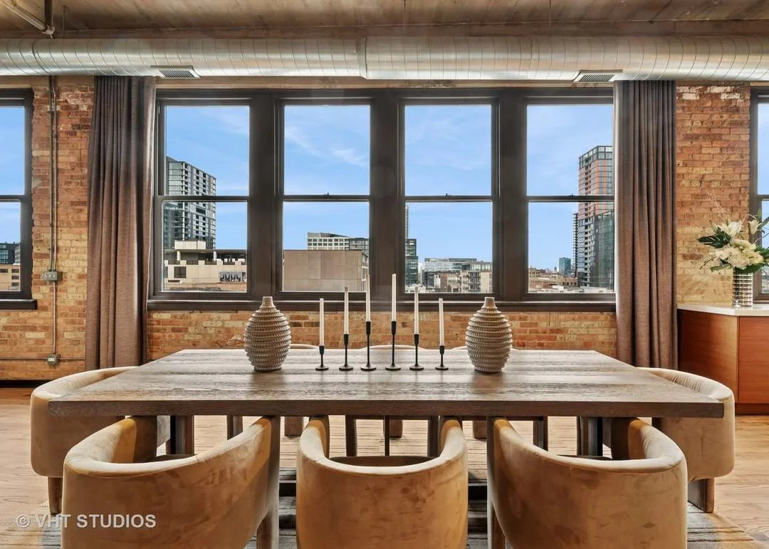 Interior of a modern dining area with a wooden table, six beige chairs, two decorative vases, and a set of white candles, with large windows showing a city skyline and blue sky.