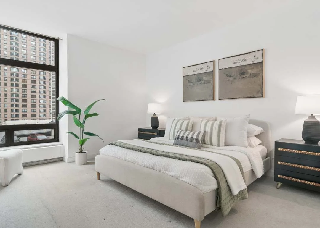 Modern bedroom with a large bed, striped pillows, two black nightstands with lamps, a tall potted plant, abstract artwork, and a window showing tall buildings outside.