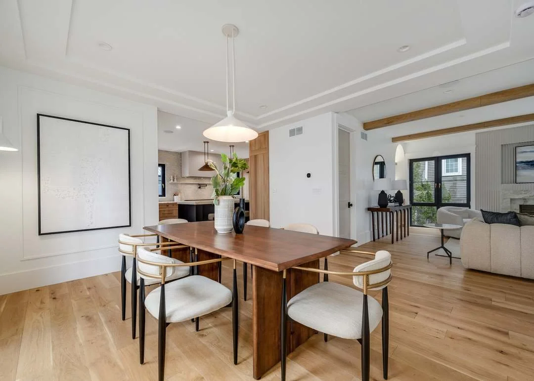 Open-concept dining and living room with wooden floors, white walls, and modern decor. The dining table is wooden with six white chairs, a white pendant light overhead, and a large, abstract artwork on the wall. The living area has a white sofa, a side table with lamps, and large windows with black frames overlooking greenery.