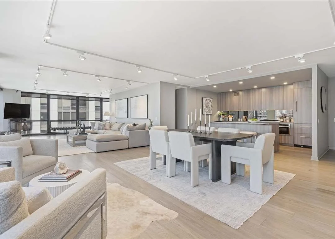 Modern open-plan living and dining room with light-colored furniture, a large window, a television, and a kitchen in the background.