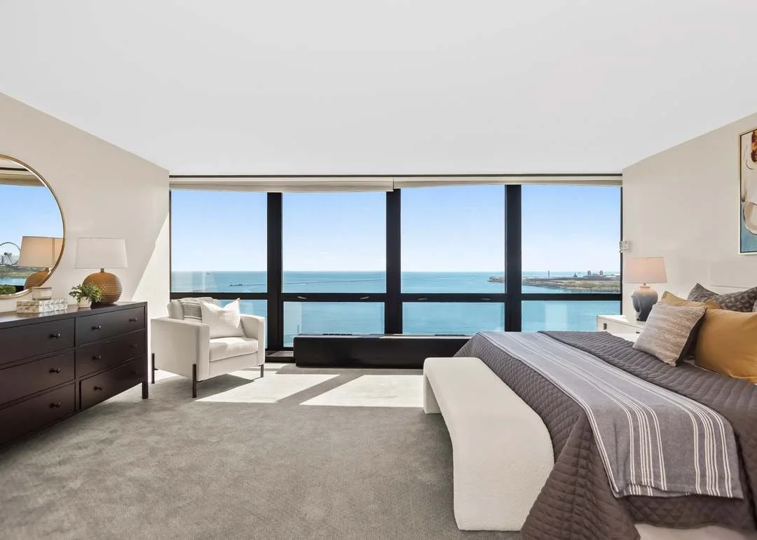 Modern hotel room with large windows overlooking the ocean, white bed with gray and beige bedding, white armchair, dark wood dresser with decorative items, and abstract artwork on the wall.