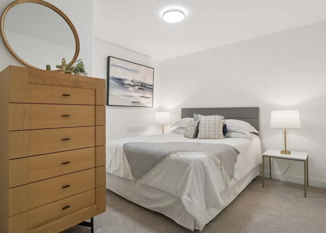 A modern bedroom with a bed, two white table lamps, framed artwork, and a wooden dresser with a round mirror above.