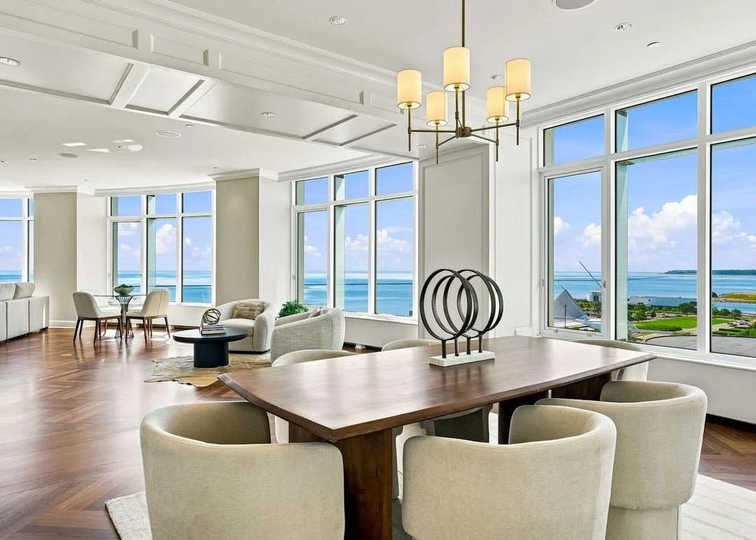 Bright, modern living room and dining area with large windows overlooking a coastal view, white walls, and wooden flooring.