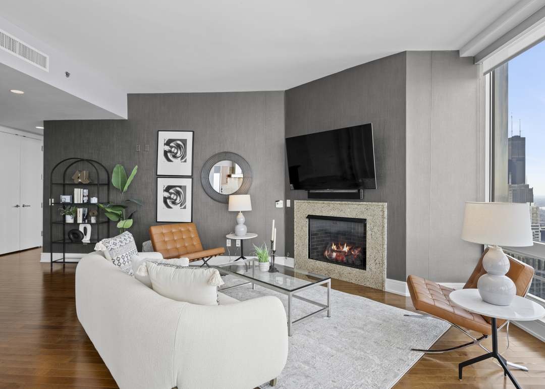 Modern living room with a white sofa, tan armchair, glass coffee table, fireplace, large flat-screen TV, large window with a city view, and decorative plants and artwork