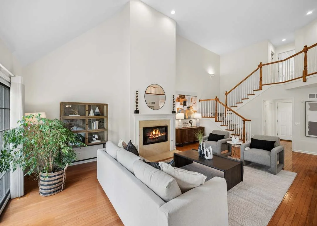 Living room with white walls, wood flooring, a fireplace, gray armchairs, a white sofa, a black coffee table, a large plant near sliding glass doors, and a staircase with wooden railing.