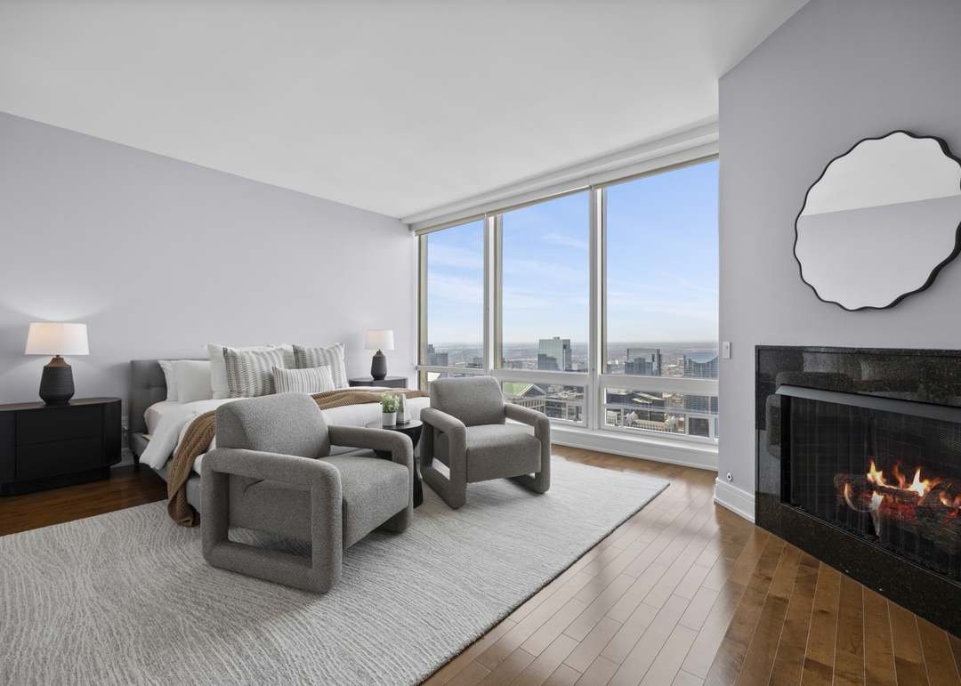 Modern living room with large windows, city view, fireplace, gray armchairs, bed, and minimalist decor.