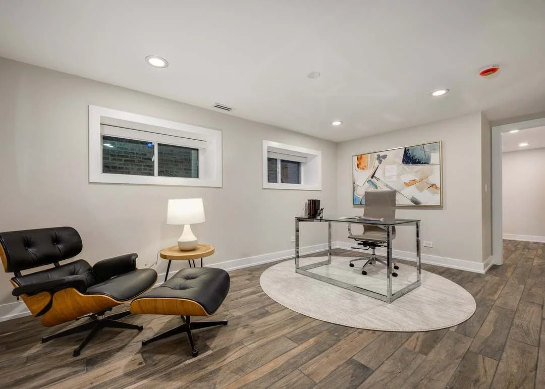 Modern home office with white walls, two small windows, a glass desk, gray swivel chair, abstract art on the wall, wood flooring, a black leather lounge chair with matching footrest, and a small side table with a white table lamp.