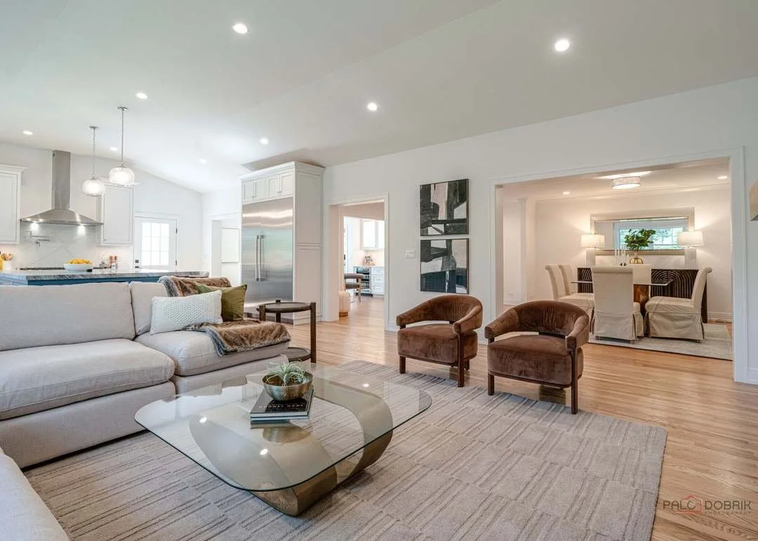 Spacious open-concept living room with cream-colored sofas, wooden chairs, a glass coffee table, and artwork on the wall. The background shows a modern kitchen with white cabinets, a stainless steel refrigerator, and a dining area with a table and chairs.