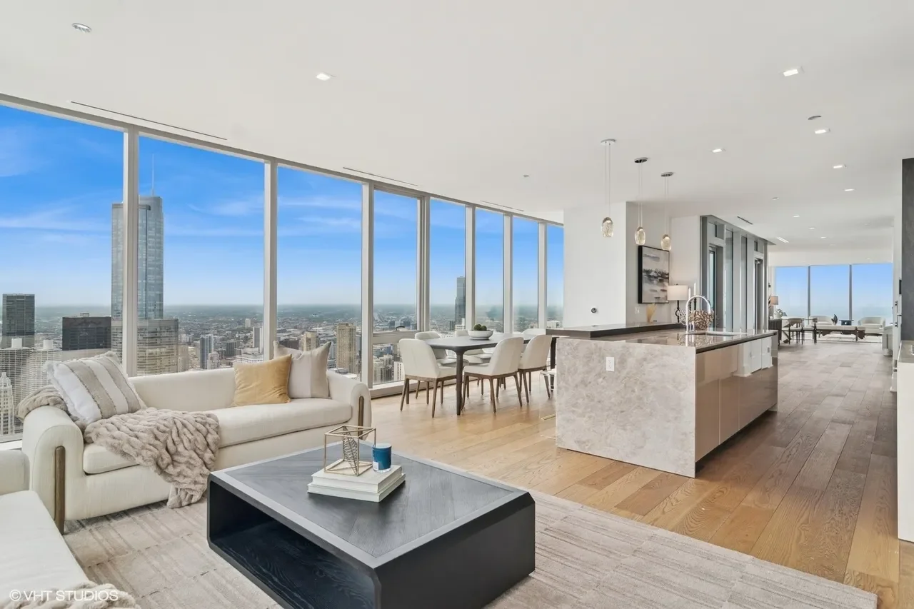 Modern high-rise apartment living room with floor-to-ceiling windows offering a city skyline view, including tall skyscrapers, white sofa with pillows and blanket, black coffee table with decorative items, dining area with white chairs and table, and a kitchen with an island, pendant lights, and hardwood floors.