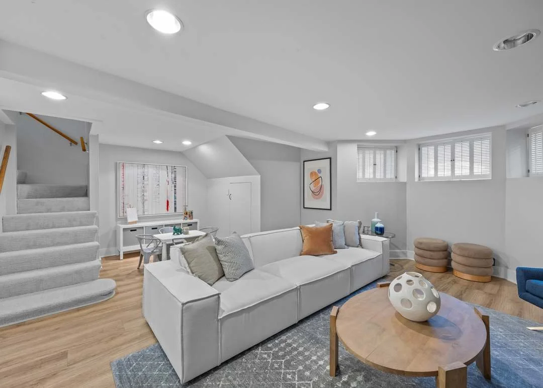A contemporary living room with a white sofa, light wood flooring, and a round wooden coffee table with a white decorative object. There's a white wall with small windows and simple artwork. A staircase with carpeted steps leads upwards on the left side.