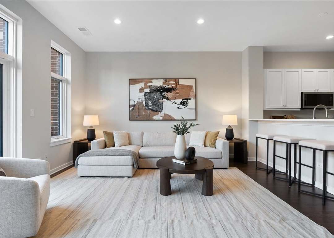 Contemporary living room with a light-colored sofa, a dark round coffee table with a white vase and decorative object, beige armchair, two side tables with lamps, abstract wall art, large windows, and an open kitchen bar area with white cabinets and barstools.