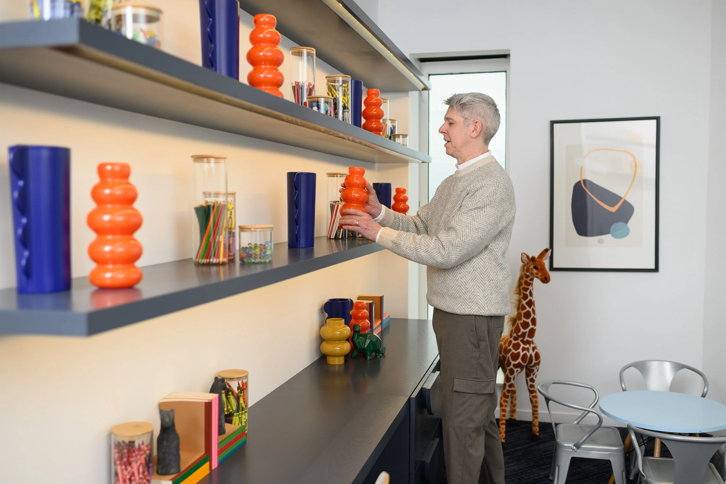 A man with gray hair and a beige sweater stands in front of a black shelf filled with colorful objects, including orange and blue vases, glass containers with art supplies, and small sculptures. A toy giraffe is visible in the background next to a framed abstract art piece, in a modern room with a window and a round dining table with chairs.