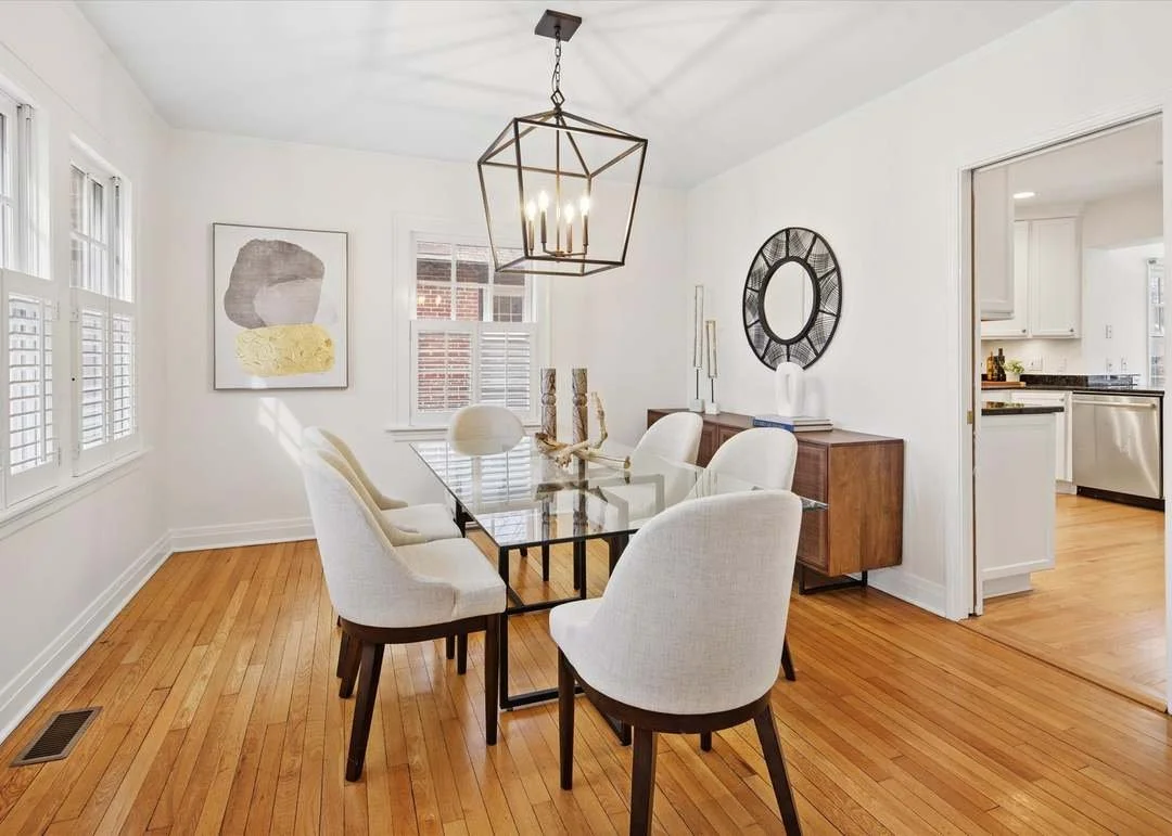 A dining room with a glass-top table surrounded by six white upholstered chairs. There is a modern geometric chandelier hanging above the table. On the wall, there are two pieces of artwork and a decorative round mirror. A wooden sideboard with decorative items and candlesticks is against the wall. To the right, an open doorway leads to a kitchen with white cabinets and stainless steel appliances. The room has light-colored walls, hardwood floors, and bright natural light from windows with white shutters.