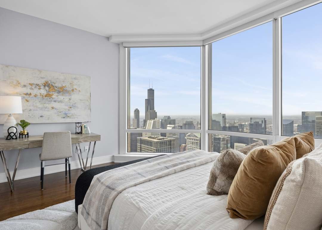 Modern bedroom with a large window view of a city skyline, including the Willis Tower, a bed with tan and beige pillows, a desk with a chair, a lamp, and wall art.