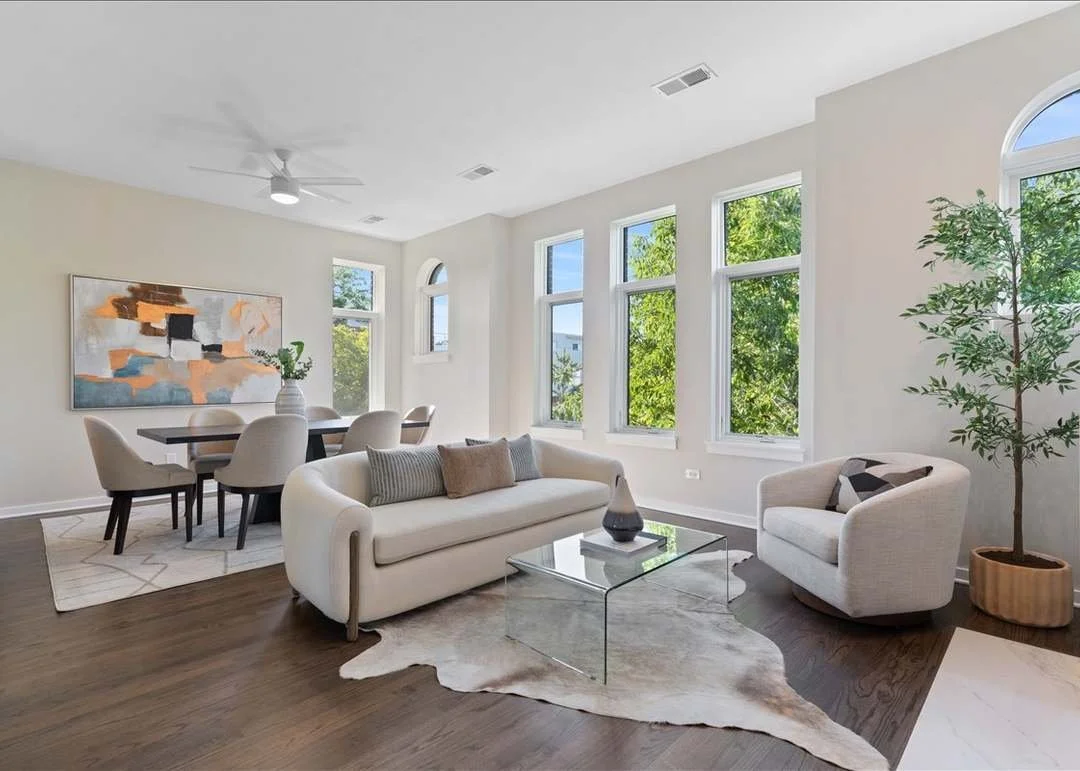 Bright living room with white walls, large windows with green trees outside, a white sofa, a glass coffee table with a decorative vase, an armchair, a potted plant, and an abstract wall art